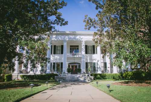 A grand white mansion with columns, surrounded by greenery and a clear blue sky. Pathway leads to the entrance.