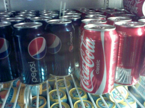 Cans of Pepsi and Coca-Cola stacked in a refrigerator, showcasing their distinct branding and colors.