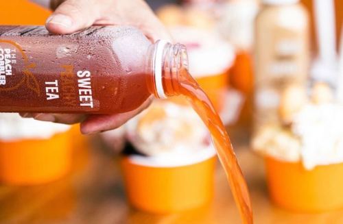 A hand pours sweet tea from a bottle into a cup, with blurred dessert cups in the background.