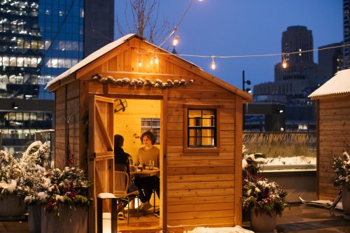 A cozy wooden cabin on a rooftop, with two people dining inside, surrounded by snow and city lights at dusk.