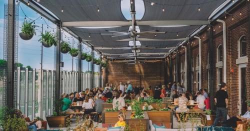 A lively rooftop venue filled with people dining and socializing under string lights and shaded canopies.