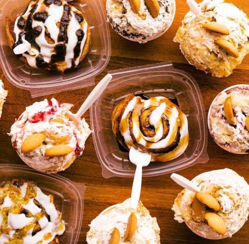A variety of desserts in clear containers, including cinnamon rolls and creamy treats, topped with cookies and drizzles.