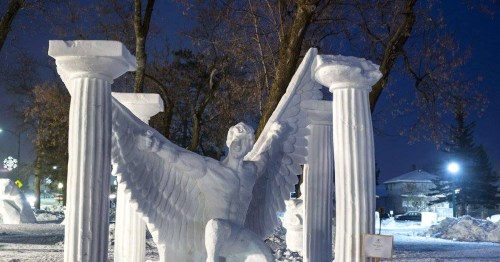 A snow sculpture of a winged figure surrounded by classical columns, illuminated at night.