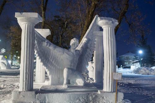 A snow sculpture of a winged figure kneeling between two columns, illuminated at night with trees in the background.