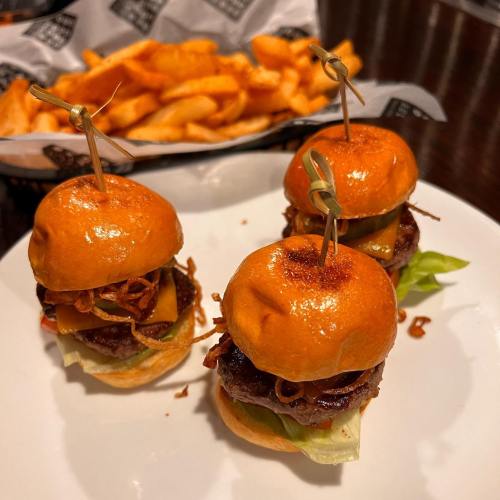 Three mini burgers with cheese, lettuce, and crispy onions on a plate, accompanied by a side of curly fries.