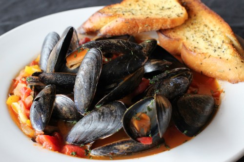 A plate of mussels in a tomato sauce, garnished with herbs, served alongside toasted garlic bread.