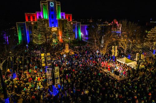 A large crowd gathers at night, enjoying festive lights and decorations around a building, with a stage and performers.