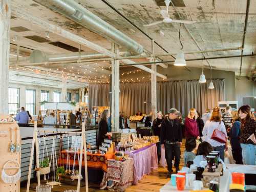 A vibrant market scene with vendors and shoppers in a spacious, well-lit venue featuring handmade goods and crafts.