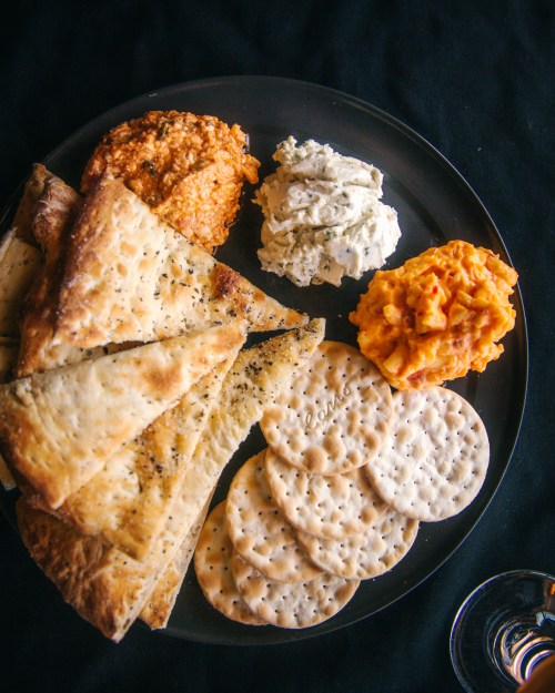 A black plate with various spreads, crackers, and bread, including hummus, cheese spread, and pita.