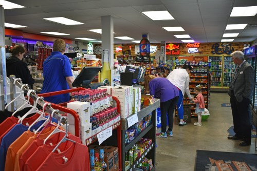 A convenience store interior with customers browsing, colorful displays, and shelves stocked with snacks and drinks.