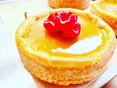 A close-up of a cheesecake topped with a raspberry and a glossy glaze, set on a light background.