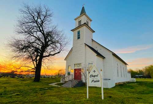 A white chapel with a steeple at sunset, surrounded by green grass and a large tree, with a sign reading "Chapel on the Prairie."