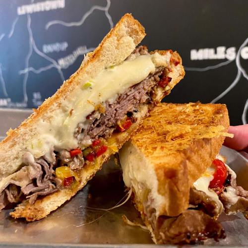 A close-up of a grilled sandwich filled with melted cheese, roast beef, and colorful peppers, held above a metal tray.