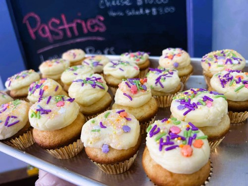 A tray of decorated cupcakes with colorful sprinkles, displayed in front of a chalkboard menu.