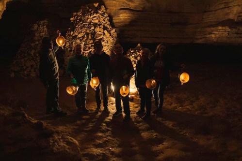 A group of six people holding lanterns in a dimly lit cave, with rock formations in the background.