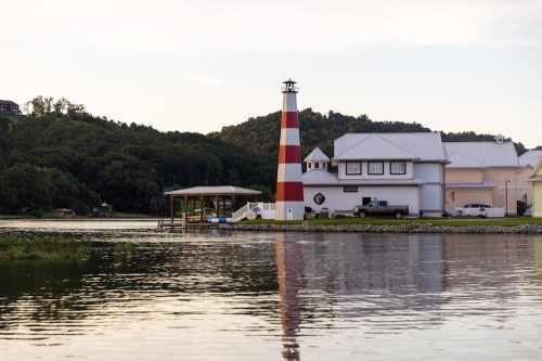 A red and white striped lighthouse beside a house on a calm lake, with trees and hills in the background.