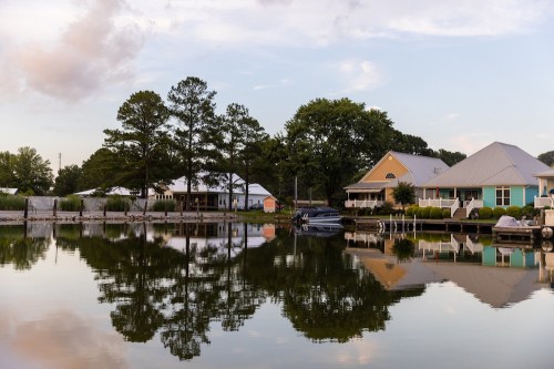 Serene lakeside scene with houses reflecting in calm water, surrounded by trees and a cloudy sky.
