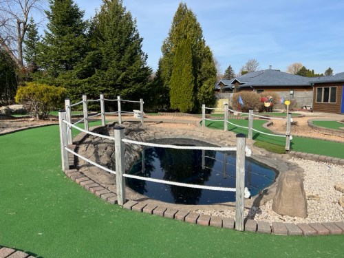 A small pond surrounded by a wooden fence, with green grass and landscaping in a mini-golf setting.
