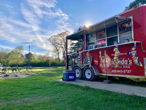 A red food truck parked on grass, with a blue sky and trees in the background, serving BBQ.