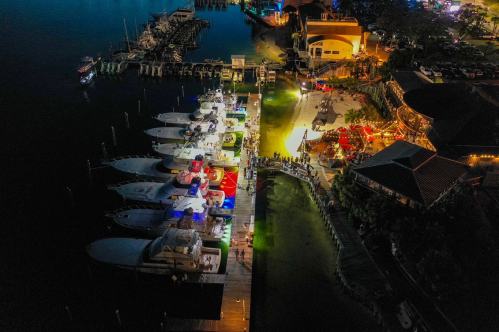 Aerial view of a marina at night, featuring illuminated boats and a lively waterfront restaurant scene.