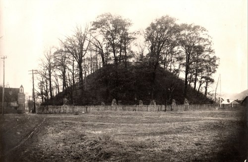 A historic burial mound surrounded by trees, with a wooden fence and a road nearby, in a sepia-toned photograph.