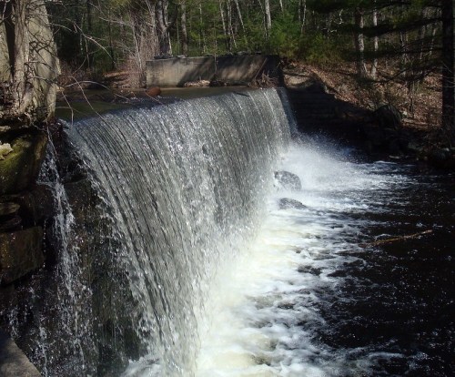 A serene waterfall cascading over a stone ledge into a dark, flowing river surrounded by trees.
