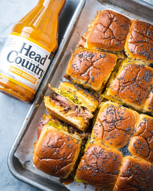 A tray of pulled pork sliders with poppy seed topping, next to a bottle of Head Country sweet mustard sauce.