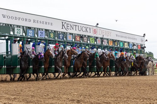 Horses burst from the starting gate at the Kentucky Derby, with colorful jockeys racing on a dirt track.