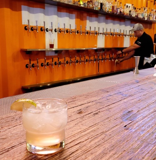 A cocktail sits on a wooden bar, with a bartender pouring drinks from a row of taps in the background.