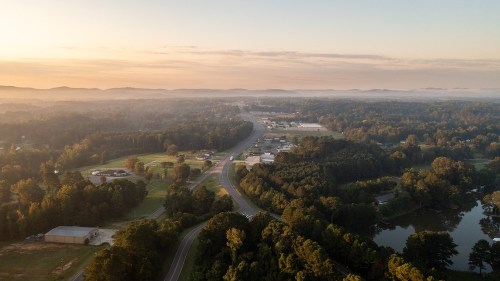 Aerial view of a small town surrounded by trees and mountains, with a winding road and misty morning light.