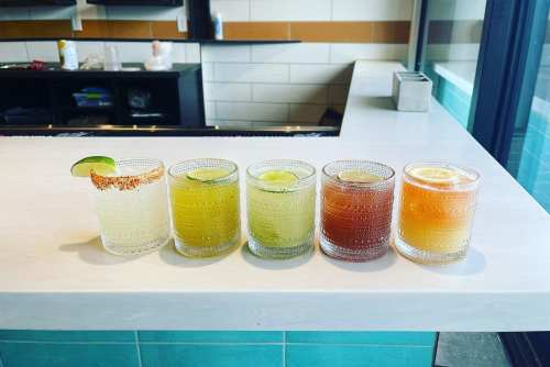 Five colorful cocktails in textured glasses lined up on a white countertop, with lime and lemon garnishes.