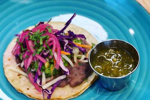 A colorful taco topped with cabbage, onions, and salsa, served on a blue plate with a side of green sauce.