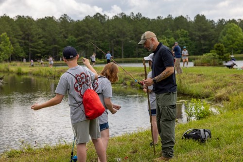 A man teaches children how to fish by a pond, surrounded by greenery and other anglers in the background.