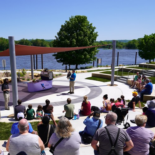 A speaker presents to a diverse audience by a river, with trees and seating areas in the background on a sunny day.
