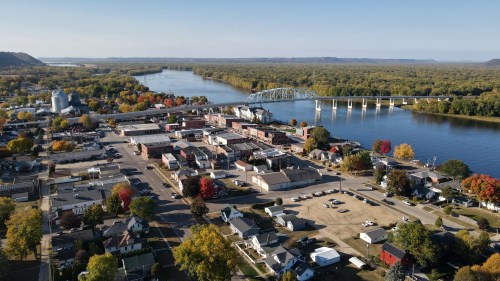 Aerial view of a small town by a river, featuring colorful autumn trees, a bridge, and a mix of buildings and homes.