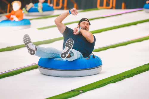 A man joyfully sliding down a snowy slope on a blue tube, with other people in the background enjoying the activity.
