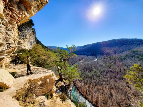 A person stands on a rocky ledge overlooking a river valley, surrounded by trees and mountains under a bright sun.