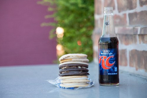 A stack of cookies next to a bottle of RC soda on a table, with greenery in the background.