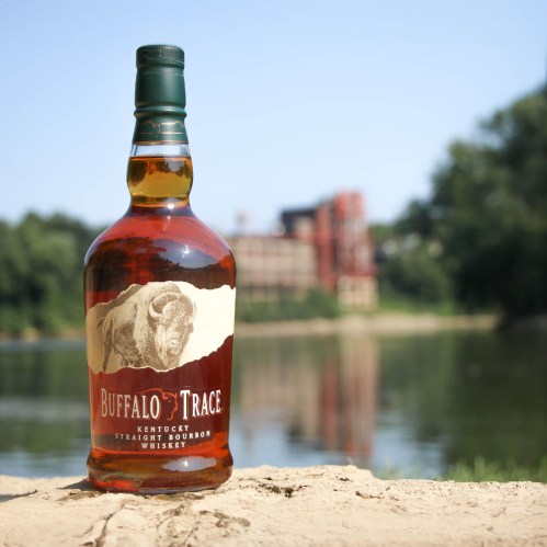 A bottle of Buffalo Trace bourbon whiskey sits on a sandy bank by a river, with a distillery in the background.