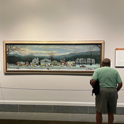 A person observes a large painting of a snowy street scene with houses and trees in a gallery setting.