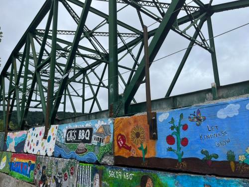Colorful murals on a concrete wall beneath a green metal bridge, featuring messages of kindness and community.