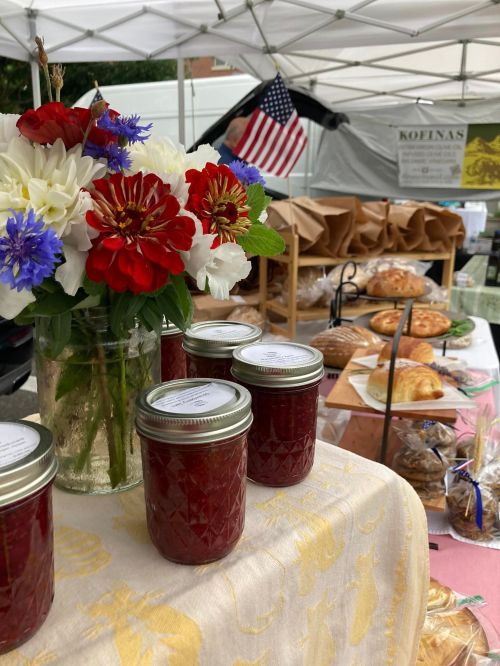 A table at a market featuring jars of jam, baked goods, and a bouquet of flowers with an American flag in the background.