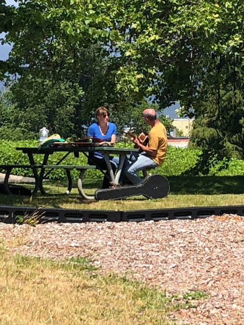A man plays guitar while a woman sits at a picnic table in a sunny park surrounded by greenery.