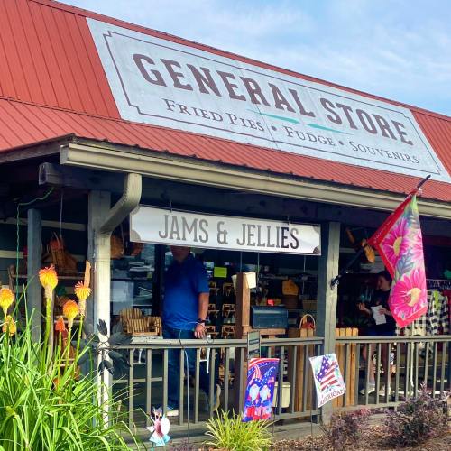 A rustic general store with a red roof, featuring a "Jams & Jellies" sign and colorful flags, surrounded by greenery.