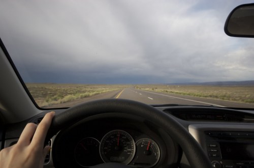 A driver's hand on the steering wheel, with a wide, open road stretching into the distance under a cloudy sky.