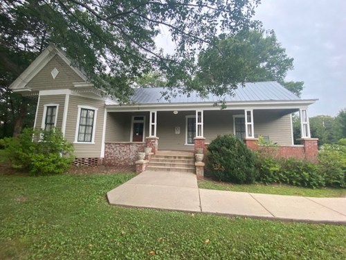 A charming gray house with a metal roof, front steps, and greenery surrounding it.