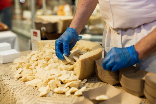 A person in blue gloves portions cheese from a large wheel onto a table covered with paper.