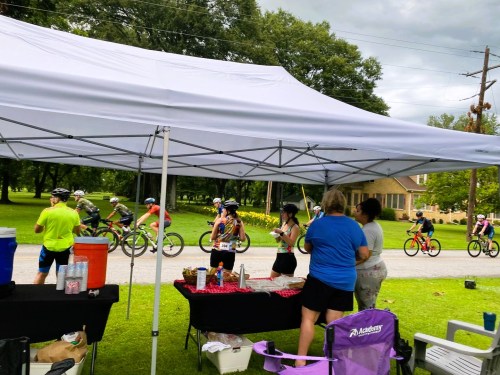 A group of cyclists passes by a tent where people are serving refreshments on a grassy area.