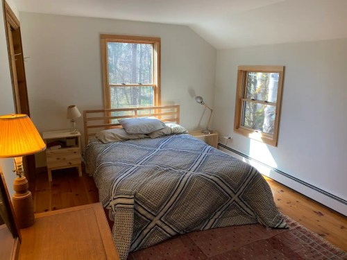 Cozy bedroom with a bed, bedside table, lamp, and windows overlooking trees. Warm, inviting atmosphere.