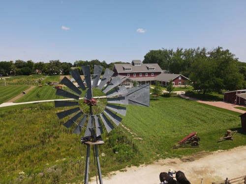 A windmill stands in the foreground with a farm and green fields in the background under a clear blue sky.
